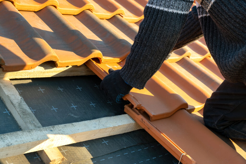 Closeup of worker hands installing yellow ceramic roofing tiles mounted on wooden boards covering residential building roof under construction.